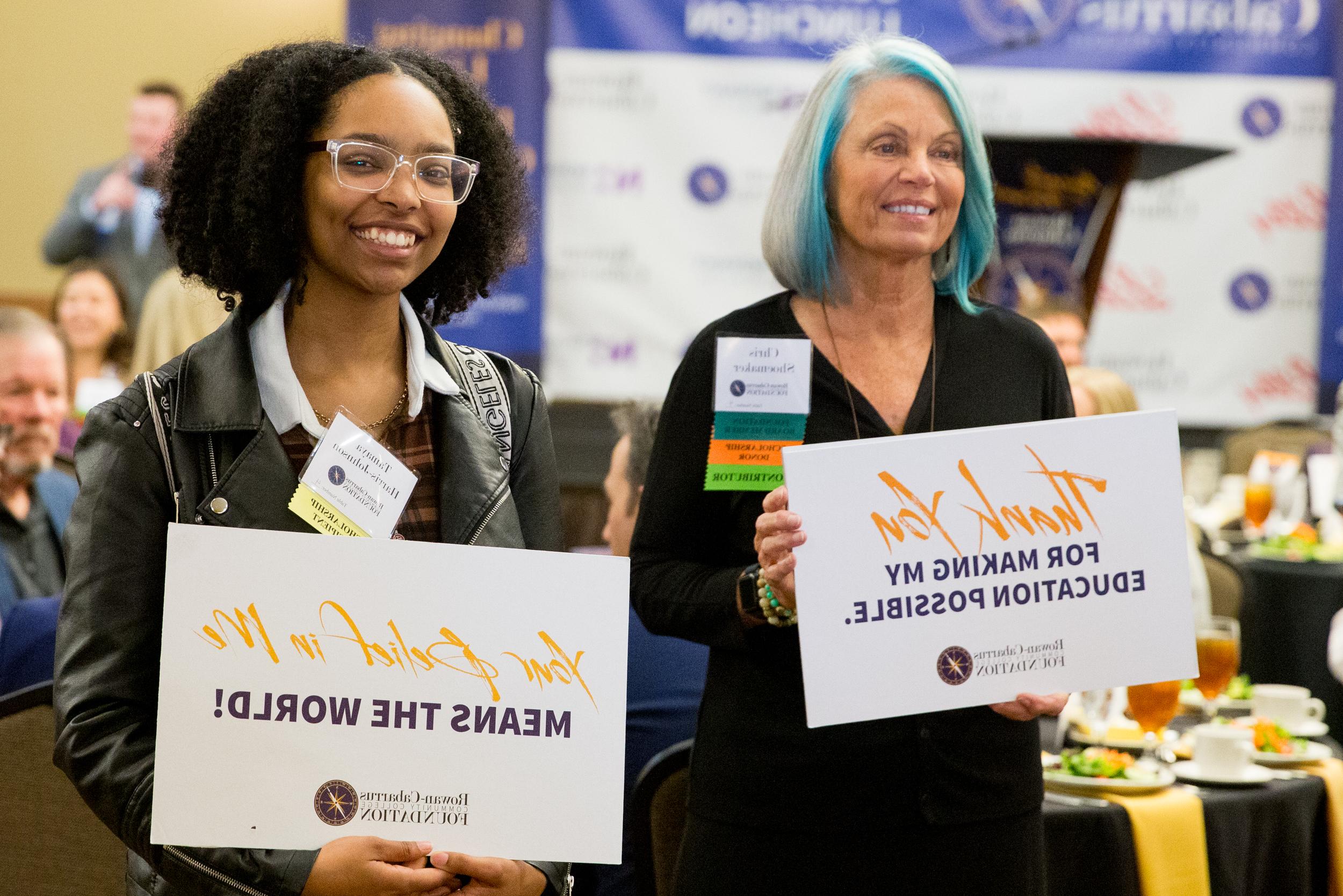 Two women hold signs at an event, thanking supporters of the Rowan-Cabarrus Foundation. Transcribed Text: Left sign: "Thank You for Making My Education Possible. Rowan-Cabarrus Foundation." Right sign: "Your Belief in Me Means the World! Rowan-Cabarrus Foundation."